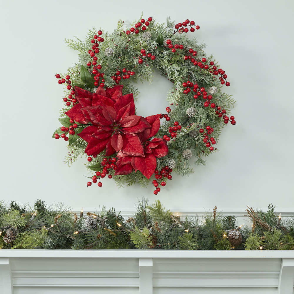 A Christmas wreath with red poinsettias and berries above a mantle covered in evergreen garland and twinkling lights. 